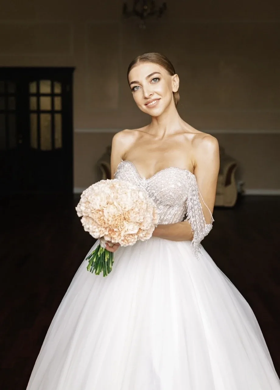 Mariée souriante tenant un bouquet pêche dans une robe de mariée perlée, maquillage et coiffure de mariée par Elena Potapenko