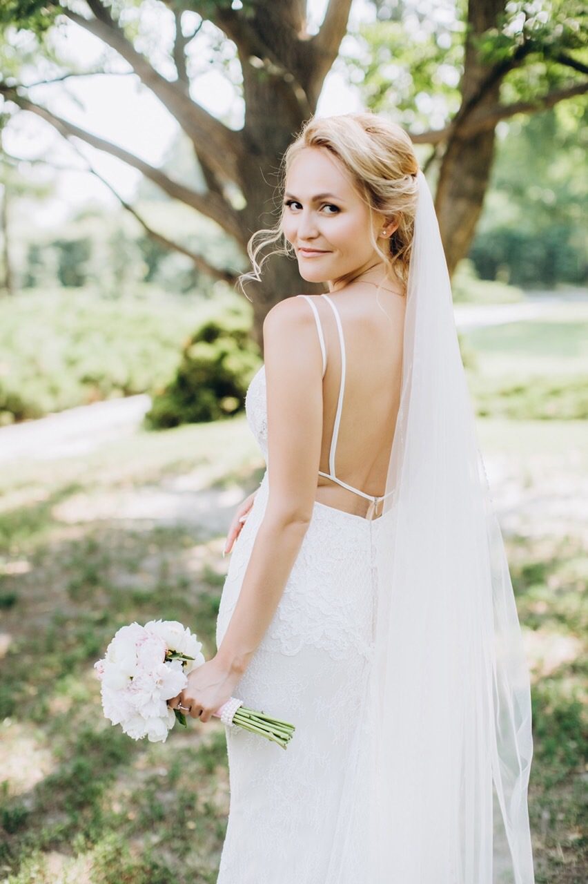Mariée blonde en robe dos nu et long voile dans un parc, regard par-dessus l'épaule