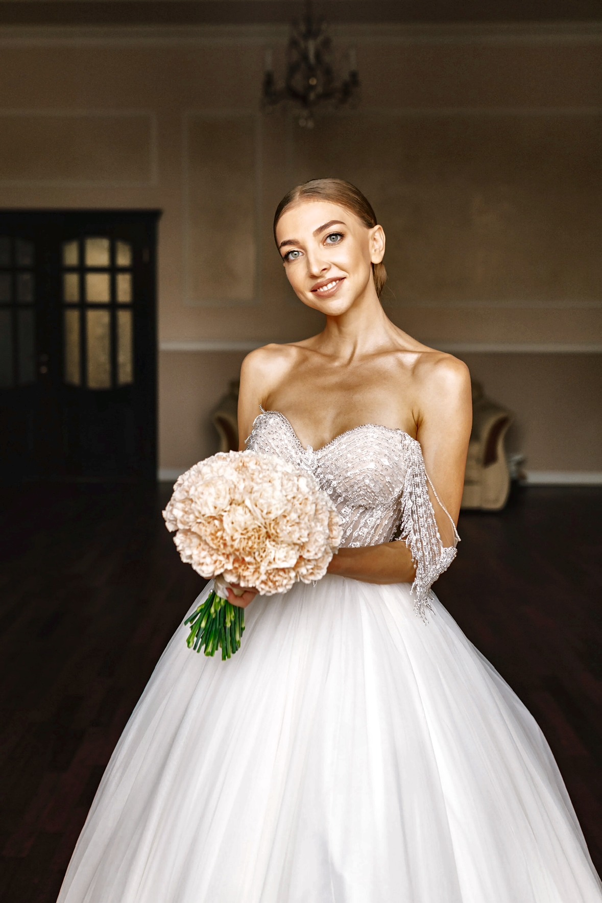Mariée souriante en robe de bal bustier perlée, tenant un bouquet pêche généreux dans une salle élégante