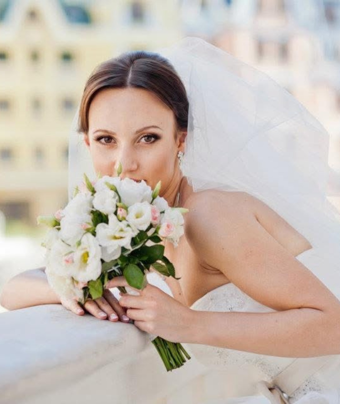 Mariée au voile flottant tenant un bouquet blanc et rose près du visage sur un balcon ensoleillé