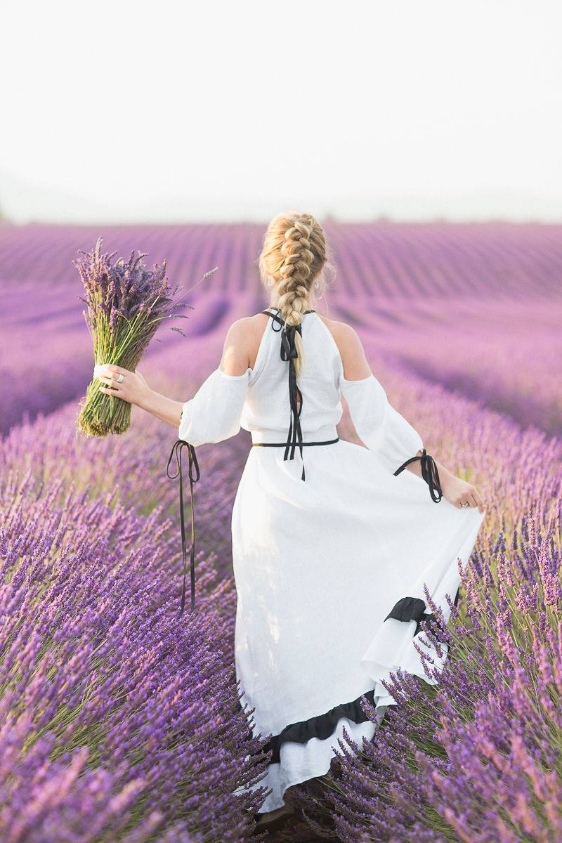 Rückansicht einer geflochtenen Hochsteckfrisur in einem Lavendelfeld, mit einem Lavendelstrauß in der Hand